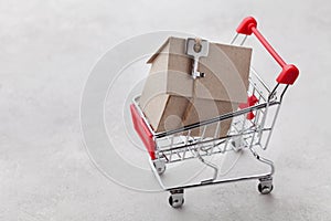 Shopping cart with model of cardboard house on gray background, buying a new home or sale of real estate concept