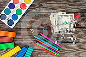 Shopping cart with money. Office and school supplies on old wooden background.