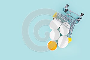 Shopping cart with raw chicken eggs and an eggshell half with yolk on a light-blue background