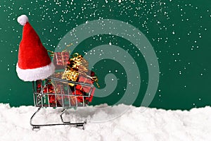 Shopping cart, santa hat, gifts on beautiful green background with snow. Front view. Christmas, new year concept. Copy