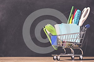 Shopping cart with school supply in front of blackboard. Back to school concept