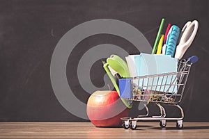 Shopping cart with school supply in front of blackboard. Back to school concept