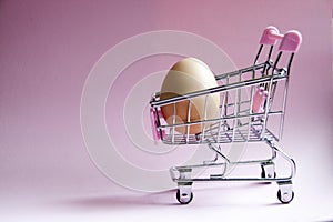 Shopping cart. Supermarket trolley with egg on pink background. Consumerism concept photo.