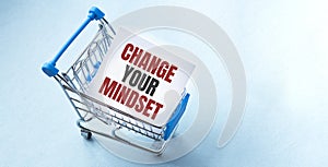 Shopping cart and text CHANGE YOUR MINDSET on white paper note list. Shopping list concept on blue background