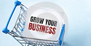 Shopping cart and text GROW YOUR BUSINESS on white paper note list. Shopping list concept on blue background