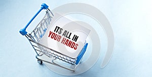 Shopping cart and text its all in your hands on white paper note list. Shopping list concept on blue background