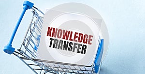 Shopping cart and text KNOWLEDGE TRANSFER on white paper note list. Shopping list concept on blue background