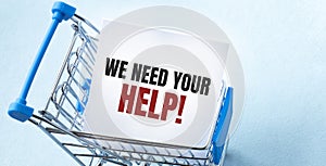 Shopping cart and text we need your help on white paper note list. Shopping list concept on blue background