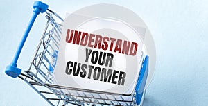 Shopping cart and text UNDERSTAND YOUR CUSTOMER on white paper note list. Shopping list concept on blue background