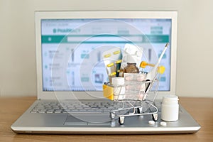 Shopping cart toy with medicaments in front of laptop screen with pharmacy web site on it. Pills, blister packs, medical bottles,