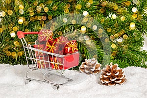 Shopping for Christmas presents. Closeup of red shopping cart and many small gifts in snow with pine cones, fir tree and bokeh