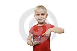 Shopping concept. Blond little smiling boy in a red T-shirt holding a small metal shopping trolley and pointing at it with his