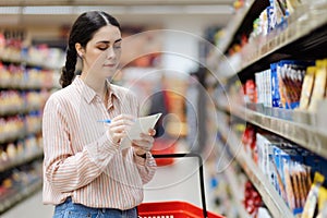 Shopping concept. Young caucasian woman choosing groceries in supermarket. Millennial young woman holding list of