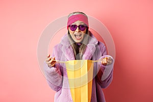 Shopping and fashion concept. Stylish asian elderly woman in sunglasses and faux fur coat open paper bag with gifts