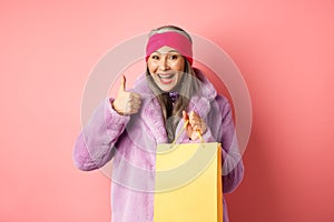 Shopping and fashion. Happy asian senior woman in stylish clothes holding paper bag from store, showing thumb-up in