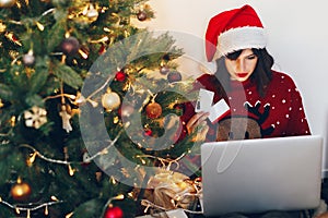 shopping online and christmas sale concept. stylish woman in reindeer hat holding credit card and laptop, under christmas tree li