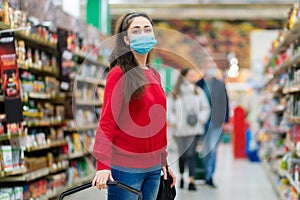 Shopping. Portrait of a young woman in a medical mask poses in the aisle of a supermarket. The concept of consumerism and the new