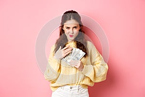 Shopping. Sassy young woman smiling and looking confident, hugging money, holding dollar bills on chest, staring