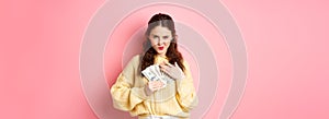 Shopping. Sassy young woman smiling and looking confident, hugging money, holding dollar bills on chest, staring