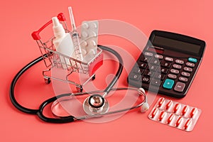 Shopping trolley cart filled pills, bottle and syringe, calculator on pink background, selective focus