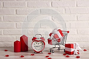 Shopping trolley with gift boxes, heart shape alarm clock and candles against the white brick wall
