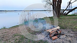 On the shore of the lake in the evening twilight after sunset, a fire of pine firewood burns. The smoke is spreading over the calm