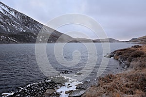 The shore of a large lake, taken by the first ice at the foot of a high snow-covered mountain, on a cloudy autumn morning