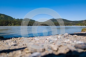 The shore of a mountain river with stones and rubble opposite which is a green forest with trees under a blue sky. Landscape on a