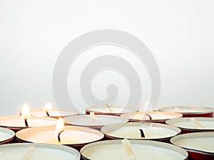 Short candles are burning against a white background. Lots of small candles. Not all candles are lit