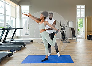 Short curly black hair coach with moustache and beard teach young woman in sportswear how to do lord of the dance yoga pose.