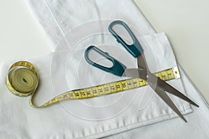 Shortening jeans. White jeans, Measuring tape, scissors on table. Jeans cutting.