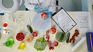 In the shot above, a scientist is sitting. Around her on the table are vegetables and laboratory supplies. Shes holding