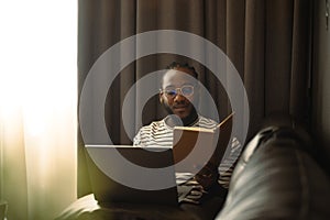 Shot of African man freelancer reading notes while working with laptop on sofa