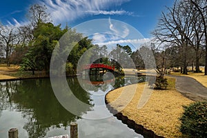 A shot of an arched red and black bridge over a lake surrounded lush green trees reflecting off the water with blue sky