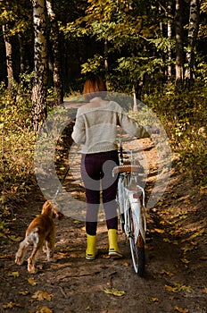 Shot from the back of a young woman walking her dog in the autumn forest and riding a bike.