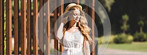 Shot of beautiful girl in white dress touching straw hat and smiling with closed eyes