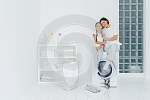 Shot of beautiful woman and his small daughter embrace and smile pleasantly, sit on washing machine, wash linen in laundry room,