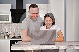 A shot of beautiful young caucasian couple standing at kitchen table in front of open laptop computer, looking at screen and