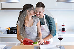 Beautiful young couple having romantic moments, hugging and kissing while cutting vegetables in the kitchen at home.