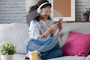 Beautiful young woman listening to music with headphones while using her smartphone sitting on sofa at home