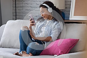 Beautiful young woman listening to music with headphones while using her smartphone sitting on sofa at home