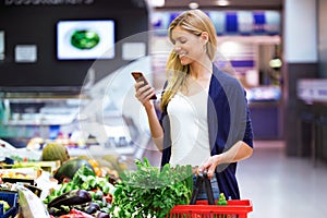 Beautiful young woman looking shopping list on mobile phone while buying fresh vegetables in the market.
