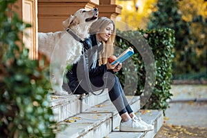 Beautiful young woman reading a book while sitting with her lovely golden retriever dog in the park in autumn