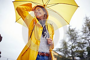 Shot from below of a young cheerful woman with a yellow raincoat and umbrella who is in a good mood while walking the city on a