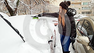 Shot of blond smiling girl in jacket clean up the snow covered car . Scraping the windshield of snow at the early snowy