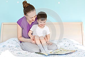 Shot of caring mother on maternity leave and small boy read fairy tale before sleep, pose at bed, view photos, enjoy domestic