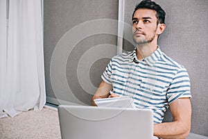 Shot of cheerful young European man in stylish shirt sitting on the floor at home with laptop pc on his lap, thinking about some