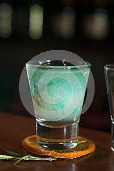 Shot glass with blue alcohol drink on dried orange slice with rosemary on wooden table