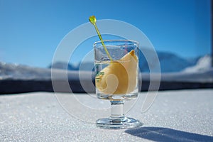 a shot glass filled with schnapps and a piece of pear stands in the snow in front of the snow covered mountains of the Alps