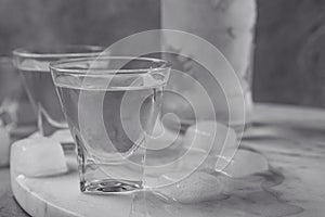 Shot glasses and bottle of vodka with ice cubes on marble board, closeup